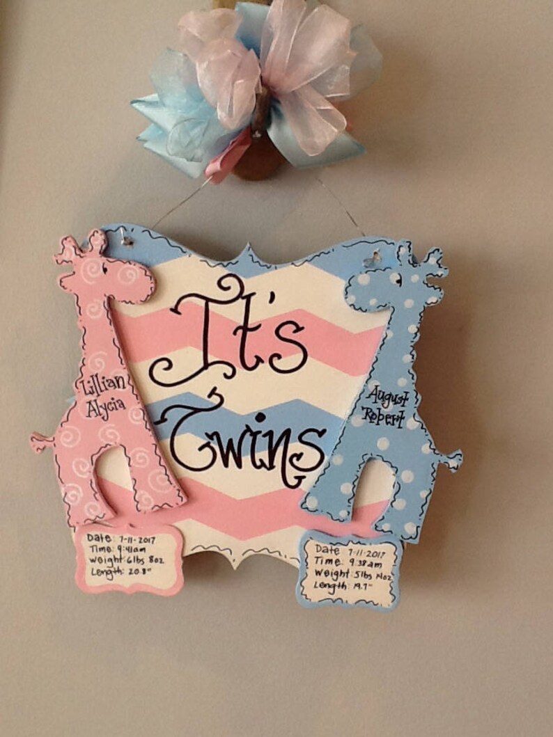 May include: A decorative wooden sign with a pink and blue chevron pattern featuring the text It's Twins. Two giraffe cutouts, one pink with swirls and one blue with dots, flank the text. Each giraffe has a plaque with birth details for Lillian Alycia and August Robert, including dates, times, weights in lbs and oz, and lengths in inches.