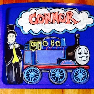May include: A blue plastic tray featuring a hand-painted illustration of Thomas the Tank Engine, the Fat Controller, and the name "Connor" in a cloud. The train is blue with red accents and smiling face.