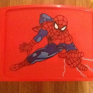 Op de afbeelding: Een felrood plastic schootbureau met een grafische illustratie van het personage Spider-Man in een dynamische houding. Het rechthoekige dienblad heeft ge&iuml;ntegreerde zijvakken en een glad oppervlak met het personage in rood en blauw afgebeeld.
