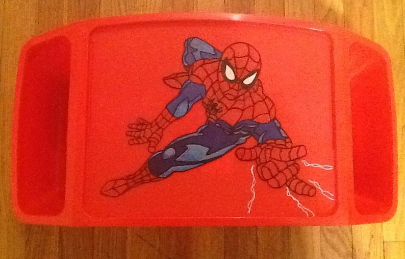 Op de afbeelding: Een felrood plastic schootbureau met een grafische illustratie van het personage Spider-Man in een dynamische houding. Het rechthoekige dienblad heeft ge&iuml;ntegreerde zijvakken en een glad oppervlak met het personage in rood en blauw afgebeeld.