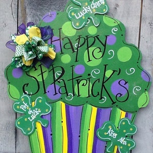 Puede incluir: Un letrero de madera con forma de cupcake en verde, morado y amarillo con el texto Happy St. Patrick's, Lucky charm, Pot of luck y Kiss Me. El letrero presenta lunares, rayas y un lazo de tela decorativo unido en el lado superior izquierdo.