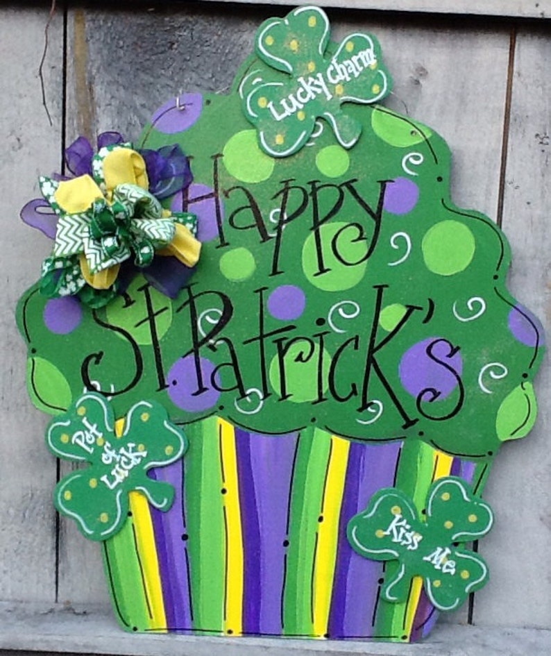 Puede incluir: Un letrero de madera con forma de cupcake en verde, morado y amarillo con el texto Happy St. Patrick's, Lucky charm, Pot of luck y Kiss Me. El letrero presenta lunares, rayas y un lazo de tela decorativo unido en el lado superior izquierdo.