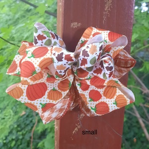May include: A decorative hair bow featuring a pattern of orange pumpkins and autumn leaves on a white background with orange polka dots, accented by brown and white gingham ribbon edges. The word small is printed in black text on the post.