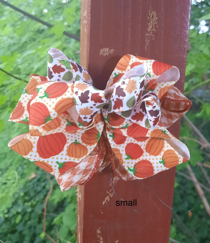 May include: A decorative hair bow featuring a pattern of orange pumpkins and autumn leaves on a white background with orange polka dots, accented by brown and white gingham ribbon edges. The word small is printed in black text on the post.