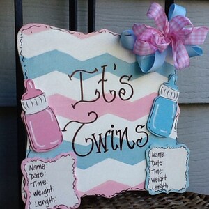 May include: A decorative wooden sign with a pink and blue chevron pattern featuring the text "It's Twins" in black script. The sign includes a pink baby bottle, a blue baby bottle, two white information plaques, and a pink and blue gingham bow.