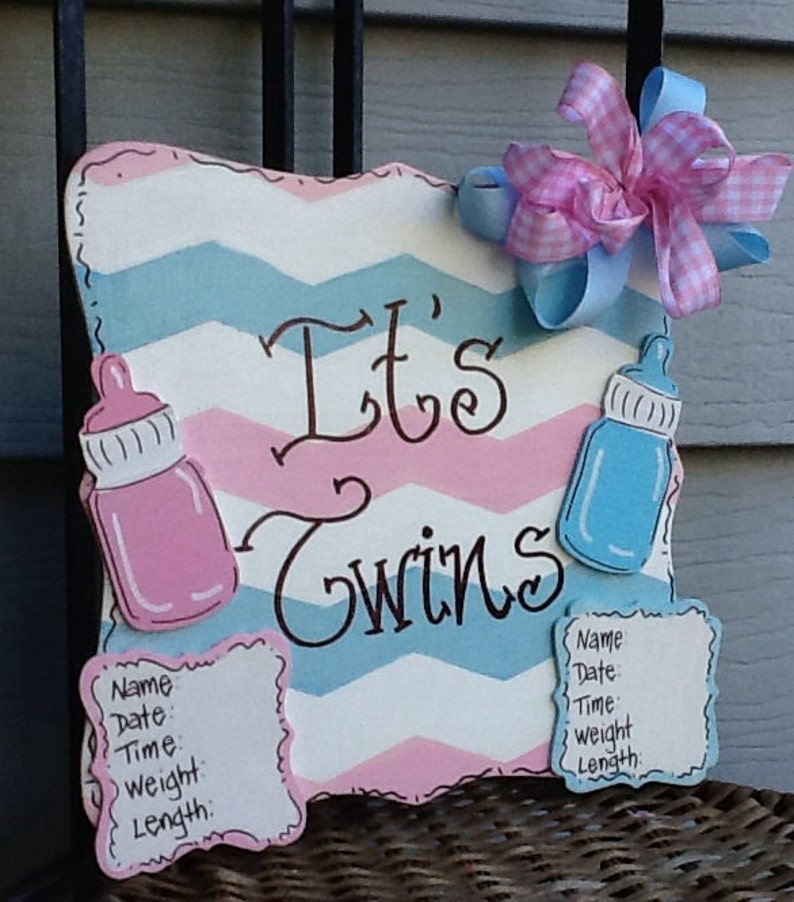May include: A decorative wooden sign with a pink and blue chevron pattern featuring the text "It's Twins" in black script. The sign includes a pink baby bottle, a blue baby bottle, two white information plaques, and a pink and blue gingham bow.