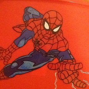 Op de afbeelding: Een rood plastic oppervlak met een grafische illustratie van het personage Spider-Man in een dynamische gehurkte houding. Het personage is afgebeeld in een rood en blauw pak met een zwart webpatroon, witte ogen en een zwart spinnenembleem op de borst.