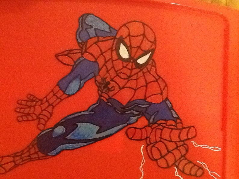 Op de afbeelding: Een rood plastic oppervlak met een grafische illustratie van het personage Spider-Man in een dynamische gehurkte houding. Het personage is afgebeeld in een rood en blauw pak met een zwart webpatroon, witte ogen en een zwart spinnenembleem op de borst.