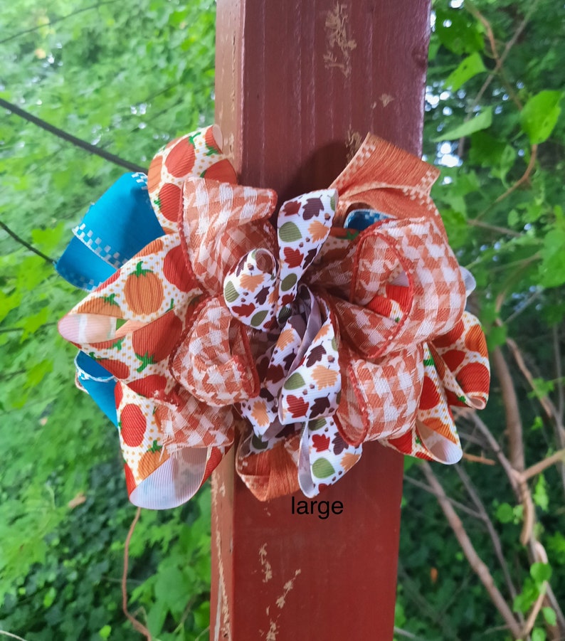 May include: A decorative bow featuring multiple ribbons with patterns of orange pumpkins, orange and white gingham, and colourful autumn leaves, accented by solid teal ribbon loops. The word large is printed in black text at the bottom.