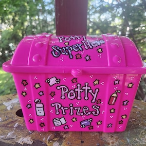 May include: A bright pink plastic treasure chest with a rounded lid. The text "Potty Super Hero" is printed on the lid, and "Potty Prizes" is on the front. The box features illustrations of toilet paper, soap, and stars in black and white.