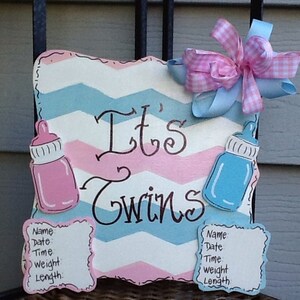 May include: A square wooden sign with a pink and blue chevron pattern featuring the text It's Twins in black script. The sign includes a pink baby bottle, a blue baby bottle, two white information plaques, and a pink and blue gingham bow.