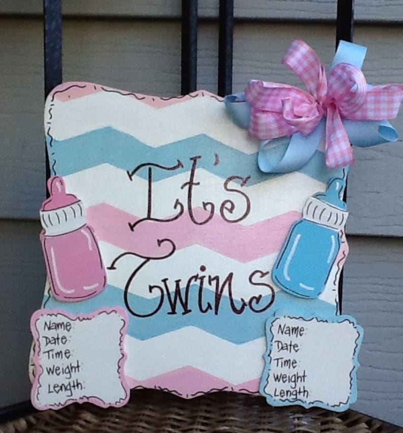 May include: A square wooden sign with a pink and blue chevron pattern featuring the text It's Twins in black script. The sign includes a pink baby bottle, a blue baby bottle, two white information plaques, and a pink and blue gingham bow.