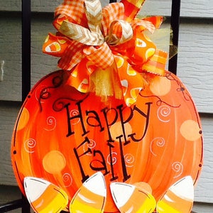 May include: A round orange pumpkin-shaped wooden sign with the text Happy Fall in black script. The sign features painted orange polka dots and swirls, four candy corn cutouts at the bottom, and a large decorative bow with orange patterns.
