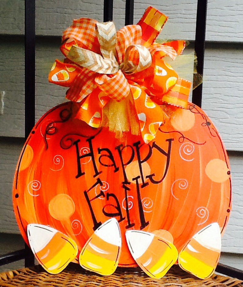 May include: A round orange pumpkin-shaped wooden sign with the text Happy Fall in black script. The sign features painted orange polka dots and swirls, four candy corn cutouts at the bottom, and a large decorative bow with orange patterns.