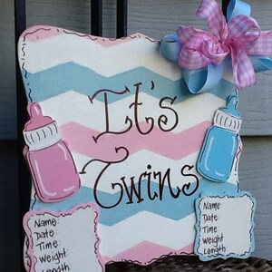 May include: A rectangular sign with a pink and blue chevron pattern featuring the text It's Twins in black script. The sign includes a pink baby bottle, a blue baby bottle, a pink and blue gingham bow, and two blank forms for birth details.