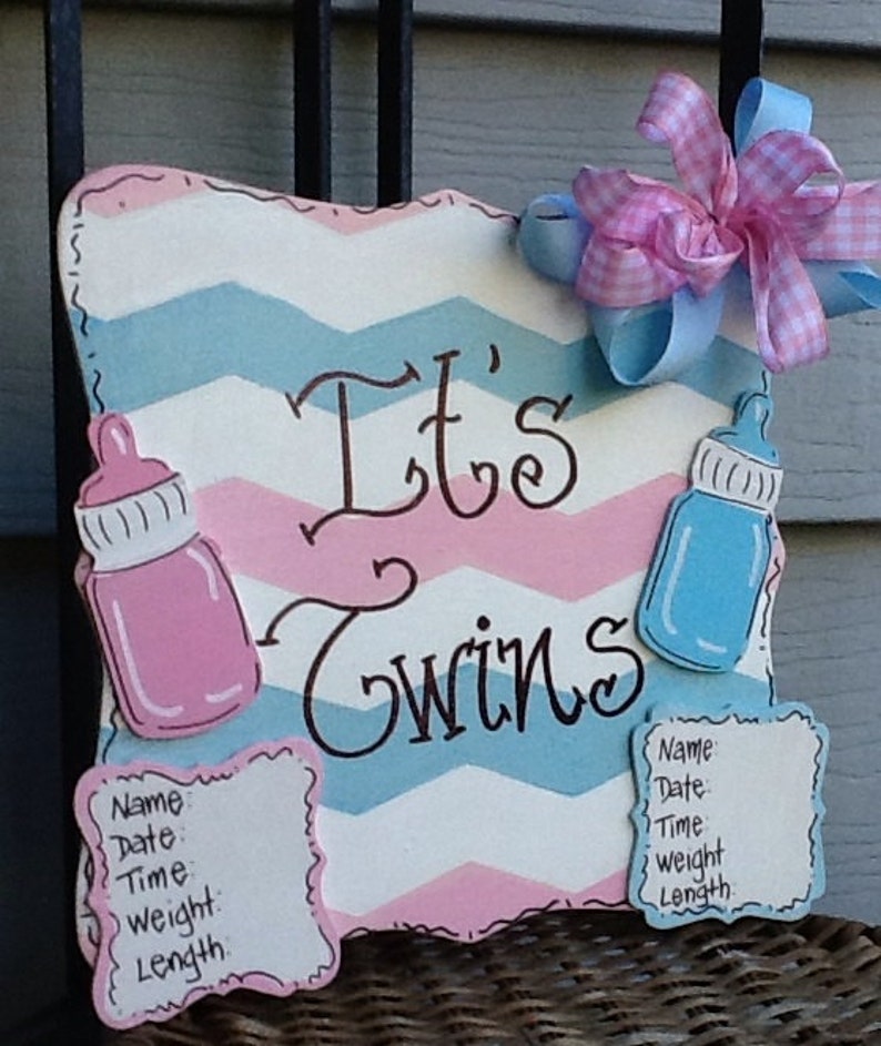 May include: A rectangular sign with a pink and blue chevron pattern featuring the text It's Twins in black script. The sign includes a pink baby bottle, a blue baby bottle, a pink and blue gingham bow, and two blank forms for birth details.