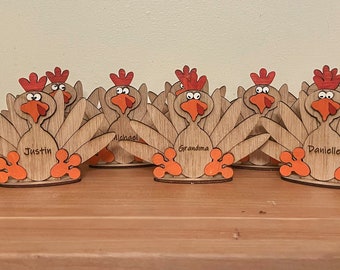 Puede incluir: Tarjeteros de pavo de madera de Acción de Gracias con nombres como "Justin", "Grandma" y "Danielle". Cada pavo tiene una cresta roja, pico y patas naranjas, y un abanico de plumas de madera. También hay una calabaza decorativa y una piña.