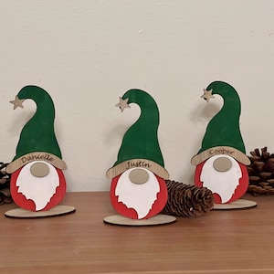 Puede incluir: Tres gnomos navideños de madera con caras pintadas de rojo y blanco, sombreros verdes y estrellas doradas. Los gnomos están personalizados con los nombres "Danielle", "Justin" y "Cooper".