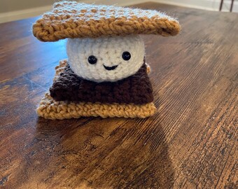 Könnte beinhalten: Ein gehäkeltes S'more mit einem lächelnden Marshmallow-Gesicht, brauner Schokolade und einem Graham-Cracker-Deckel.