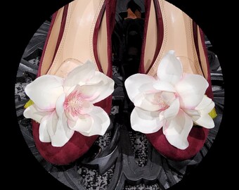 May include: A pair of burgundy suede pumps adorned with large, white and pink magnolia flower shoe clips. The shoes are set against a dark, patterned surface, showcasing the floral embellishments.
