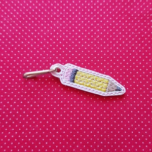 May include: A yellow pencil keychain with a silver metal clasp. The pencil is embroidered on a white fabric.