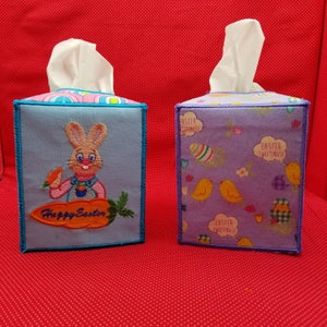 Könnte beinhalten: Zwei Taschentuchbox-Hüllen zum Thema Ostern. Eine ist hellblau mit einem Hasen, der eine Karotte hält, und dem Schriftzug "Happy Easter". Die andere ist lila mit gelben Küken und Osterei-Designs. Beide haben weiße Taschentücher, die oben herausragen.
