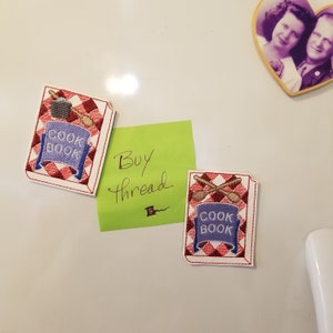 May include: Two embroidered patches with a red and white checkered background. Each patch features a blue book with the text "COOK BOOK" and a pair of wooden spoons. The patches are attached to a white surface.