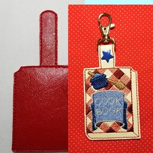 May include: Red leather key fob with a stitched loop and a red and white checkered design. The fob has a blue star and the words "Cook Book" on it.