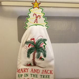May include: White kitchen towel with an embroidered palm tree with red and white striped candy canes hanging from the branches. The towel is hanging from a white and green embroidered Christmas tree towel holder. The text "Mary and Jack Up in the Tree" is embroidered at the bottom of the towel.