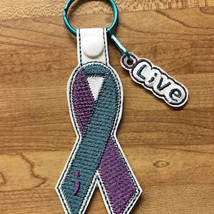 Puede incluir: Llavero de cinta bordada en color turquesa y morado con borde blanco y anilla turquesa. El diseño de la cinta incluye un punto y coma y la palabra "Live" en un dije separado.
