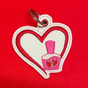 Puede incluir: Un llavero de corazón blanco con un contorno de corazón rojo. Dentro del corazón hay una botella de esmalte de uñas rosa con dos corazones rojos en la parte delantera.