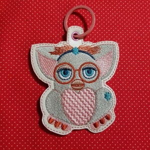 May include: A white, grey and pink embroidered keychain featuring a cartoon character with blue eyes and glasses. The character is wearing a pink headband with a blue flower.