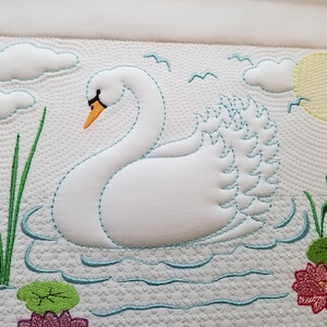 Pode incluir: Cisne branco bordado em um tecido branco com água azul, juncos verdes e nenúfares rosa. O cisne tem um bico laranja e um sol amarelo está no fundo.