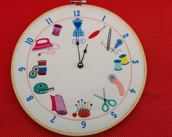 May include: A white clock face with black hands and blue numbers. The clock face is decorated with embroidered images of sewing supplies, including a sewing machine, scissors, spools of thread, and a thimble.