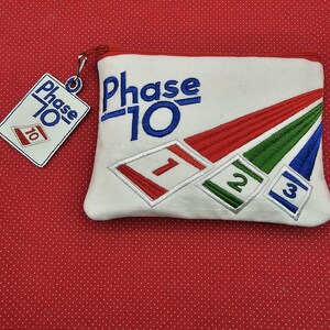 May include: White zippered pouch with the text "Phase 10" embroidered in blue. The pouch has three embroidered cards with the numbers 1, 2, and 3.