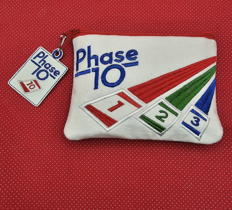 May include: White zippered pouch with the text "Phase 10" embroidered in blue. The pouch has three embroidered cards with the numbers 1, 2, and 3.