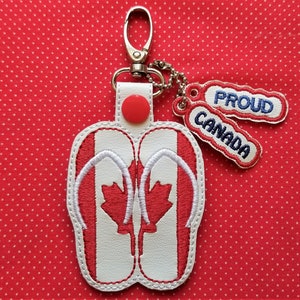 Puede incluir: Un llavero de chanclas con bandera canadiense roja y blanca, un bot&oacute;n rojo y dos etiquetas que dicen "Orgulloso" y "Canad&aacute;".