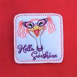 Pode incluir: Etiqueta bordada com uma avestruz de desenho animado usando óculos rosa e um colar de pérolas. A avestruz tem cílios rosa e um laço rosa no cabelo. O texto "Hello Sunshine" é bordado abaixo da avestruz.