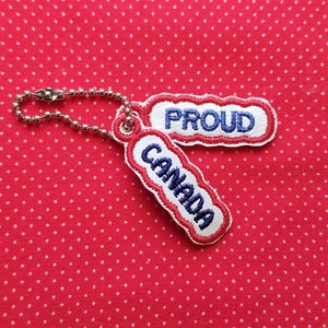 Puede incluir: Un llavero rojo y blanco con una cadena plateada. El llavero tiene dos etiquetas, una que dice "CANADA" y la otra que dice "PROUD".
