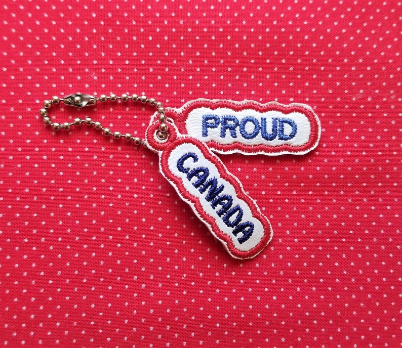 Puede incluir: Un llavero rojo y blanco con una cadena plateada. El llavero tiene dos etiquetas, una que dice "CANADA" y la otra que dice "PROUD".