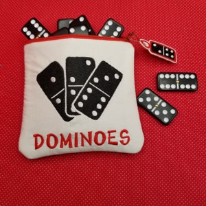 Peut inclure: Une pochette en tissu blanc avec une fermeture éclair rouge et un texte brodé en rouge qui dit "DOMINOES". La pochette est remplie de dominos noirs et il y a trois dominos noirs sur un fond rouge à pois blancs.