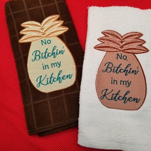 Könnte beinhalten: Zwei Küchentücher mit gestickten Ananas-Motiven. Die Ananas auf dem braunen Handtuch ist beige mit braunen Blättern und dem Text "No Bitchin' in my Kitchen" in Türkis. Die Ananas auf dem weißen Handtuch ist rosa mit braunen Blättern und dem Text "No Bitchin' in my Kitchen" in Türkis.