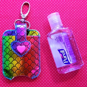 May include: A rainbow mermaid-scale patterned hand sanitizer holder with a pink heart and a silver keychain. The holder is next to a clear plastic bottle of Purell Advanced Hand Sanitizer with infused essential oils.