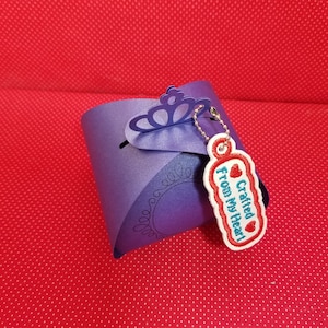 May include: A purple gift box with a decorative crown-shaped closure. A tag with the words "Crafted From My Heart" is attached. The box and tag are on a red dotted background.