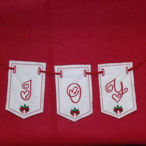May include: A white felt banner with red string, spelling out the word "JOY" in cursive. Each letter is on a separate white felt flag with a red bow and green leaves at the bottom.