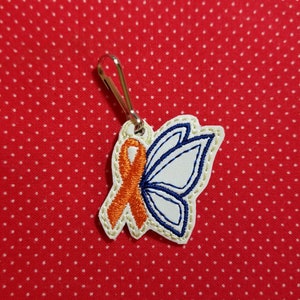 Może przedstawiać: Biały urok w kształcie motyla z pomarańczową wstążką, przymocowany do srebrnego suwaka zamka błyskawicznego. Urok jest wyszyty niebieską i białą nicią.