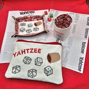 May include: A white Yahtzee game pouch with red stitching and embroidered dice and a cup. The pouch has the word "Yahtzee" embroidered in red letters. The pouch is lying on a red surface with white polka dots.