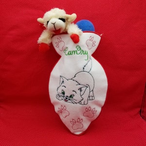 Puede incluir: Un cordero de peluche blanco con detalles rojos asomando de una bolsa de tela blanca con un gato bordado. La bolsa tiene el texto "LamDry" bordado.