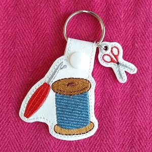 Puede incluir: Llavero de cuero blanco con una aguja de coser roja y plateada, una bobina de hilo azul y marrón y un par de tijeras rojas y plateadas.