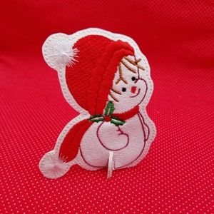 May include: White felt snowman with a red hat and scarf, embroidered with holly and a smiling face. The snowman is standing on a red background.